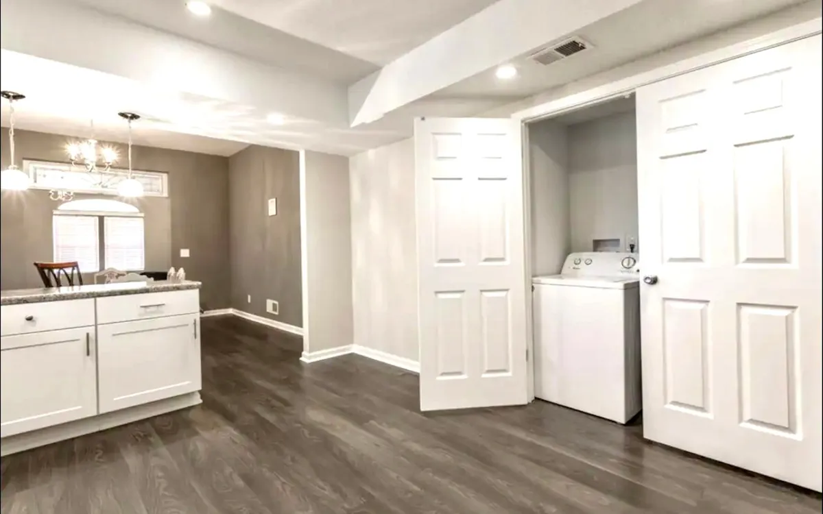 A bright, modern kitchen with a large island at the center, stylish chandelier lighting, and a washer and dryer tucked conveniently to one side. Sleek countertops and clean lines create a welcoming, practical environment ideal for preparing meals or getting ready for a day of business meetings.