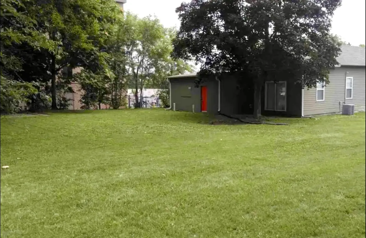 A bright green lawn leads up to a house with a cheerful orange door, flanked by trees. The setting captures an inviting, homey atmosphere perfect for outdoor dining, casual gatherings, or enjoying quiet moments. The vibrant colors and ample space emphasize comfort and openness.