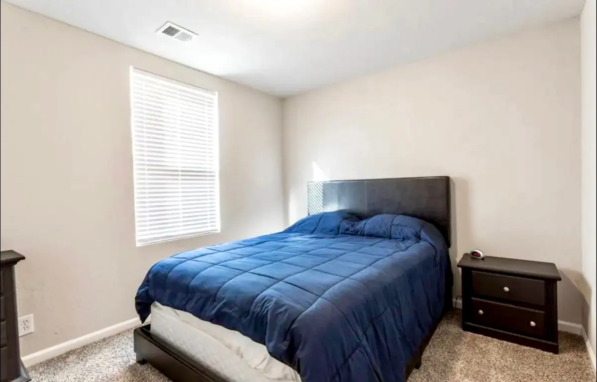 A tranquil bedroom featuring a neatly made bed with soft white linens, a nightstand with a lamp, and a window letting in natural light. The color palette is soothing, with neutral tones and minimal decor, creating a peaceful retreat for relaxation or reading.