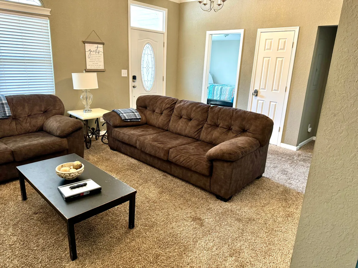 A welcoming living area with plush sofas arranged around a coffee table, with the front door visible, suggesting easy flow and openness. The space is styled for both relaxation and entertaining guests, with cozy decor.