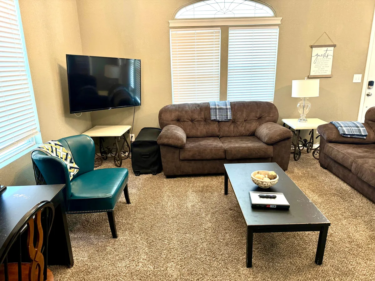A cozy, stylish living room featuring a brown sofa, blue accent chair, black coffee table, television, and warm lighting from lamps. The space looks comfortable and perfect for relaxing or socializing, with modern touches and inviting decor.
